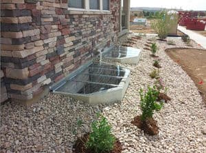 Basement window wells with stone landscaping and plants beside a residential building.