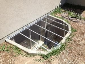 Exterior view of a basement window well covered with a transparent enclosure and surrounded by grass.