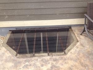 Glass basement window well cover with steel bars, surrounded by a concrete patio.