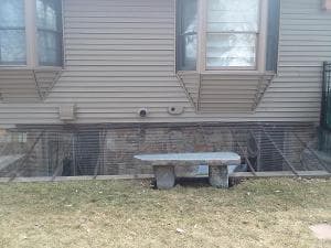 Stone bench in front of a house with window wells and brick foundation.