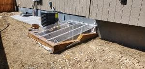 Wooden DIY greenhouse frame with plastic cover beside a house on gravel ground.