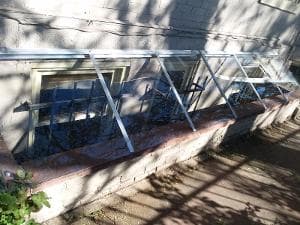 Greenhouse window structure with metal frames, showcasing plants on a sunny sidewalk.