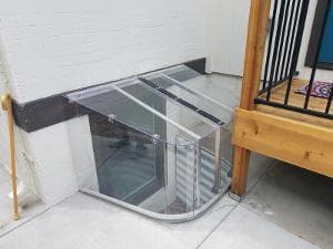 Clear acrylic basement window well cover installed beside wooden deck. Enhances safety and visibility.
