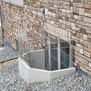 Clear basement window well cover against a stone wall with gravel landscaping.